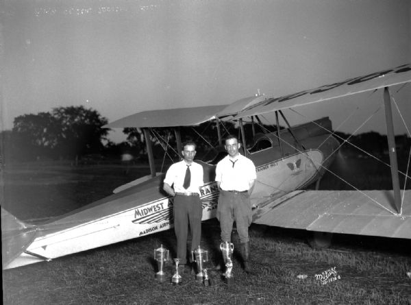William McDonald and Merl Buck posing in front of a small airplane with several Air Show trophies on the ground in front of them.