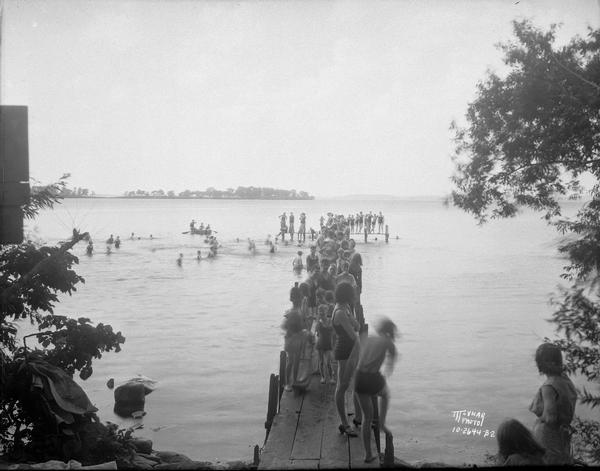 View from shoreline looking down towards bathers at the Willows Beach, standing on a pier and swimming in the lake.