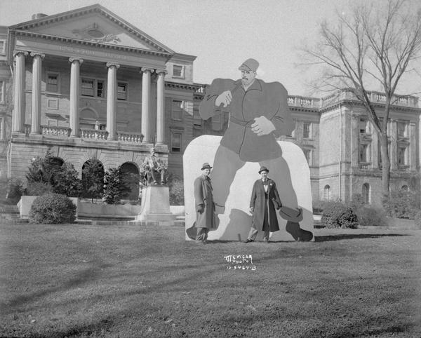 A Paul Bunyan figure used to celebrate University of Wisconsin Homecoming, was placed on the lawn in front of Bascom Hall. Two athletes are standing beside the figure, Edward Christianson, football player, and Gordon Fuller, basketball star. This effigy was later replaced by Paul Bunyan's Ax Trophy in 1948.