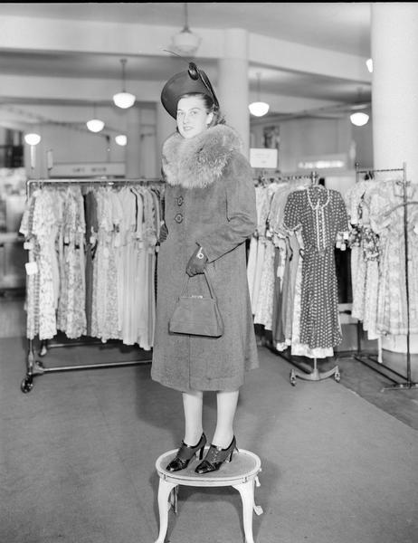Model standing on step stool wearing hat and gloves, with fur collar coat, dress shoes, and carrying a purse. Part of the fashion series from Kessenich's Ready to Wear, 201-203 State Street, showing racks of dresses.