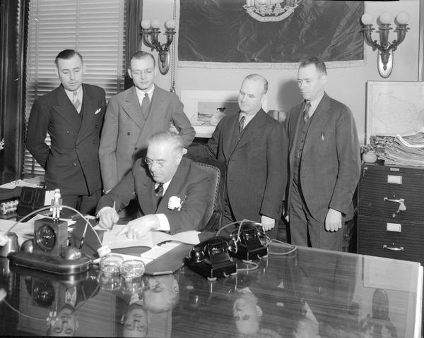 Governor Julius P. Heil sitting at his desk signing a bill, with four men looking on from behind.