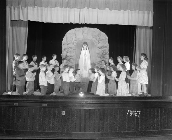Cast of Holy Redeemer School play on stage. Children are standing and kneeling in prayer in front of a shrine containing a student dressed as the Virgin Mary.