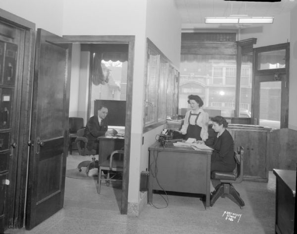 Madison News Agency, 446 West Gilman Street, office interior with room on left showing a man at a desk, and the room on the right showing two women at a desk with an adding machine.