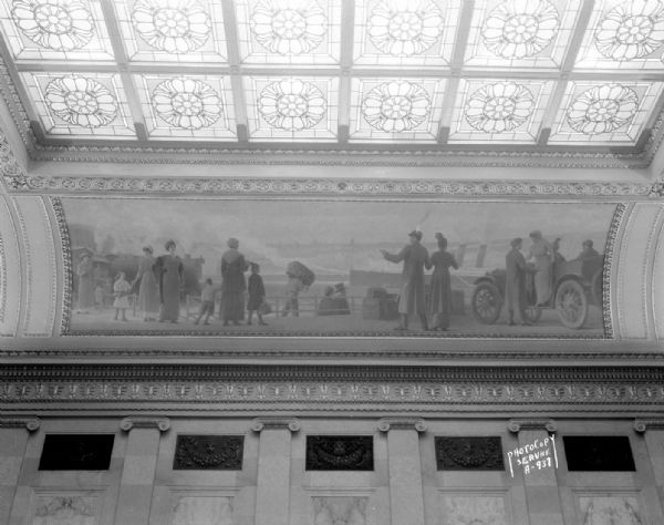 The mural depicts a 1915 harbor scene with a steamship, railway train, automobile, and airplane. One of four murals by Charles Yardley Turner, representing the evolution of transportation in America. The stained glass skylight in the Wisconsin State Capitol North Hearing Room can also be seen. The title of the mural is: "A Modern Transportation System."