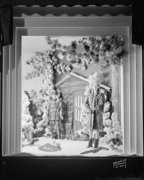 Manchester's, Inc., fur coat window display showing two mannequins, one wearing a fur coat and the other wearing a coonskin cap, standing in the snow in front of a log cabin, taken from Mifflin Street.