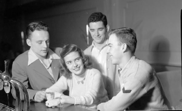 Teenage girl with three young men, one in uniform, at a soda fountain.  The boys are admiring her necklace and bracelet.