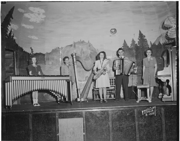 Three adult and two child musicians on stage with xylophone, harp, saxophone, accordion and piano at Smitty's Hall, for the grand opening program sponsored by the Milwaukee Cheese Company which opened a new cheese plant on April 1st.