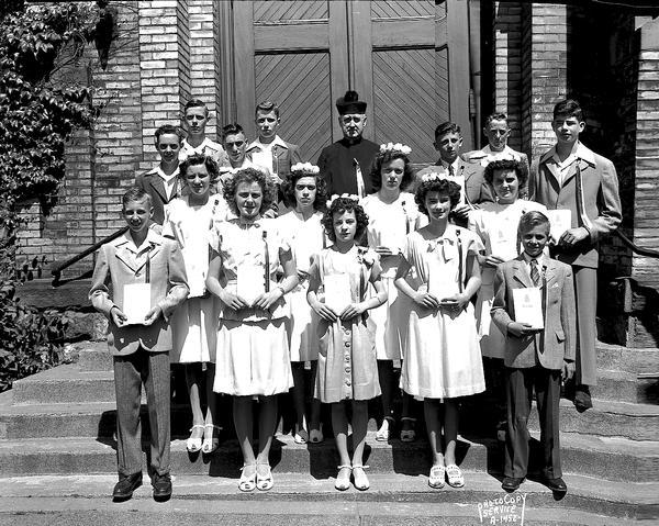 Outdoor group portrait of the graduation class of St. Patrick's School, taken on front steps of St. Patrick's Church, 410 East Main Street. Includes Reverend Albert V. Grace, pastor, nine boys, and seven girls.
