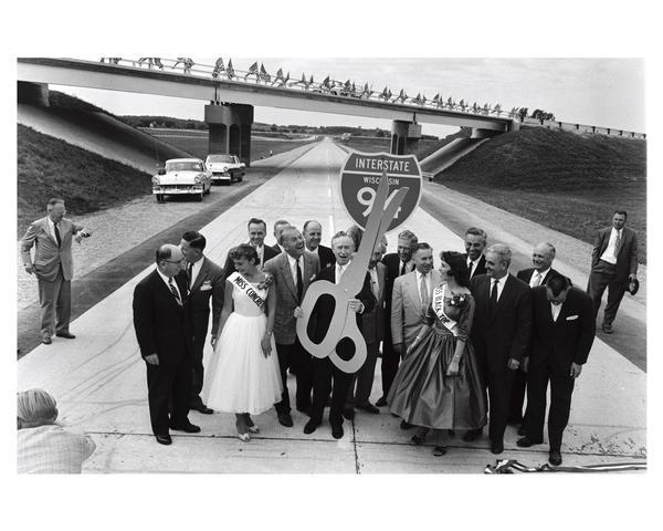 Dedication of Wisconsin's first expressway, a seven-mile stretch of Interstate Highway I-94 in Waukesha County, Wisconsin. Presiding at the ribbon-cutting ceremony are several government officials and Miss Concrete and Miss Black Top. Holding the large pair of scissors is Governor Vernon Thomson and to his right is Secretary of State Robert Zimmerman. Warren Knowles, who would later become governor, is also in the crowd.