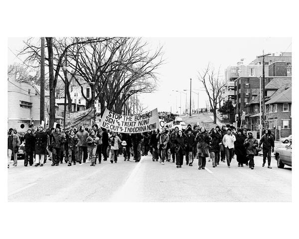 A crowd of anti-Vietnam war demonstrators march down Wisconsin Avenue with signs and banners. One banner reads: "Stop the Bombing. Sign the Treaty Now! U.S. Out of Indo-China Now!".