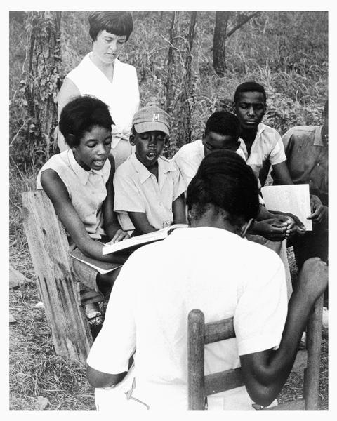 A group of African American students participating in a Freedom School class outdoors during Freedom Summer. The class, which was held on Tougaloo College campus, was led by Joanne Gavin, who is standing behind the students. Seated on the far left is Hymethia Washington, who is holding a book. Charles, who is wearing a baseball cap, is sitting next to Hymethia.