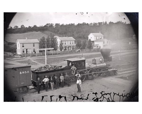 Elevated view of several men, some of whom may be railroad workers, posing in front of a Minnesota and St. Paul wood-burning engine at a train crossing. The town post office and other structures are behind the train. Station master, Samuel Barber, is fifth from left.