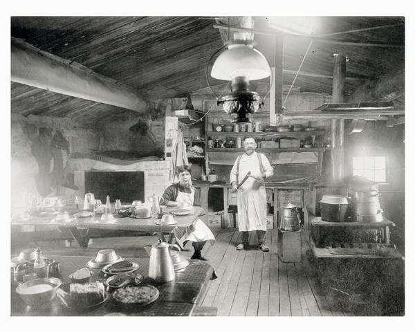 Two men preparing a meal in a lumber camp shanty or bunkhouse.