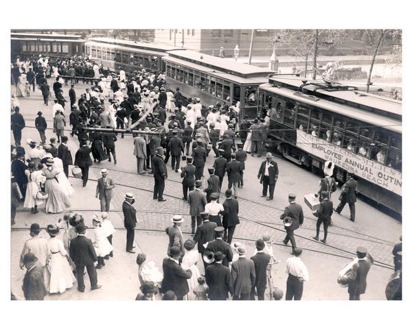 In a festive mood, Milwaukee Electric Railway and Light Company employees disembark from flag-decked trolley cars to enjoy a company-sponsored outing. The company hired trolley cars to Waukesha Beach. Recreation included picnicking, ball games, and potato races.