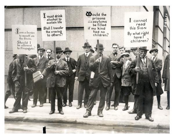 Four impoverished men hired by "The Medical Review of Reviews" carry signs with the eugenics slogans, "I am a burden to myself and the State. Should I be allowed to propagate?," "I must drink alcohol to sustain life. Shall I transfer the craving to others?," "Would prisons and asylums be filled if my kind had no children?," and "I cannot read this Sign. By what right have I children?"