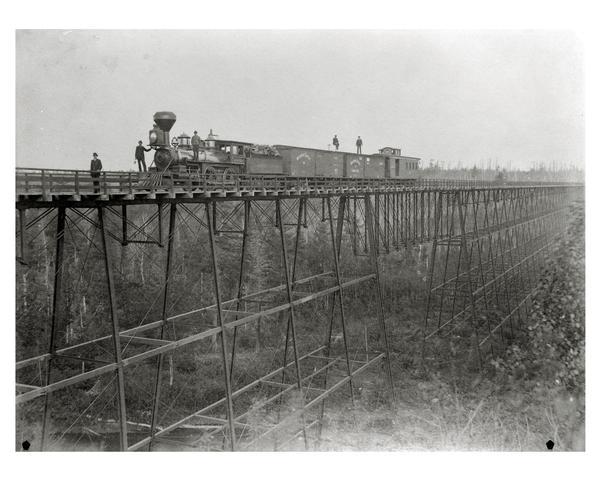 Wisconsin Central Railroad train on White River Bridge, with crew posing on top of cars. The bridge was 1600 feet long and 110 feet above the water. In 1877 excursions were run from the South so the sight-seers might view its "marvel of engineering." Bridge was dismantled and sold for junk in 1909.