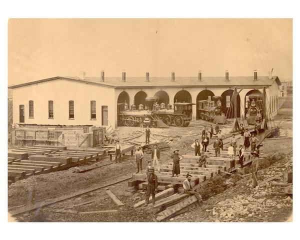 "The West Wisconsin Railway roundhouse at Eau Claire, ca. 1872. It was located about a mile north of the present Civic Center. Workmen in the foreground are building a new turntable to replace the earlier one, often call the "gallows." Locomotives in the stalls are Baldwin wood burners, all 4-4-0s, built in 1871. Headed out of stall 7, to the left, is engine No. 12 the "Geo. W. Clinton"; the engine headed into stall 6, with its tender missing and the main rods down, is engine No. 14, names for "Matt W. Carpenter"; and backed into stall 3 is engine No. 10, named the "D.A. Baldwin." Baldwin was one of the incorporators in 1871, of the St. Paul, Stillwater & Taylors Falls Railway. These engines all carried their same numbers even when they were taken over a few years later by the Chicago, St. Paul, Minneapolis & Omaha in 1878."