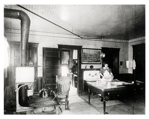 Woman baking in a large kitchen with lean-to pantry and a wood-burning stove. The sconces or reflectors on the kerosene lamps intensified the light.