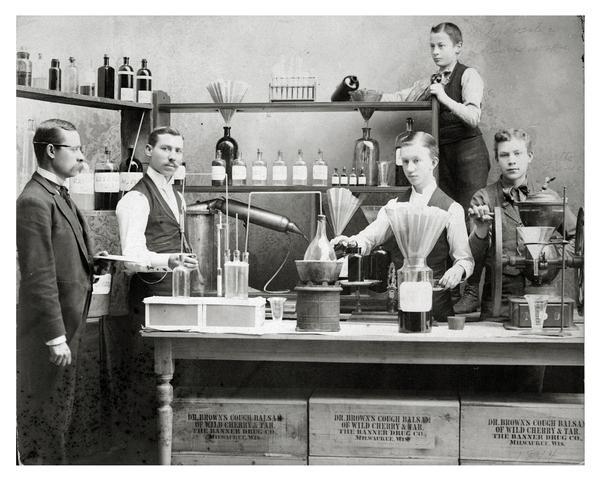 Workers posing in front of pharmaceutical equipment to demonstrate the making of Dr. Brown's Cough Balsam at Kienth Drugs and Medicines, 608 Mitchell Street. The original caption reads: "Dad and Uncle Frank in the Lab making Dr. Brown's Cough Balsam they invented." On the front of the photograph individuals are identified as follows, from left to right:  Uncle Frank, Dad, unidentified, Sylvester Kurzewski, Otto Frank.