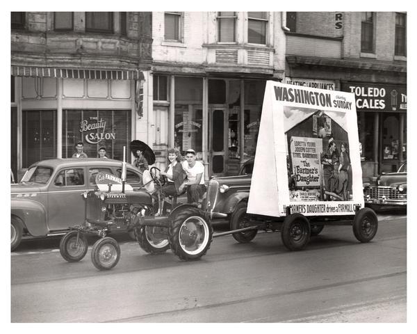 Two people driving a Farmall Cub through an urban area. They are towing a wagon with movie advertisement, announcing "The Farmer's Daughter," starring Loretta Young, Joseph Cotton, and Ethel Barrymore. The advertisement also proclaims that the Farmer's Daughter Drives a Farmall Cub.