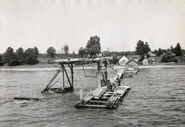 View from water of fishing dock and structure alongside; log buildings in background on shore. Possibly on shoreline of one of the Apostle Islands or on Chequamegon Bay.