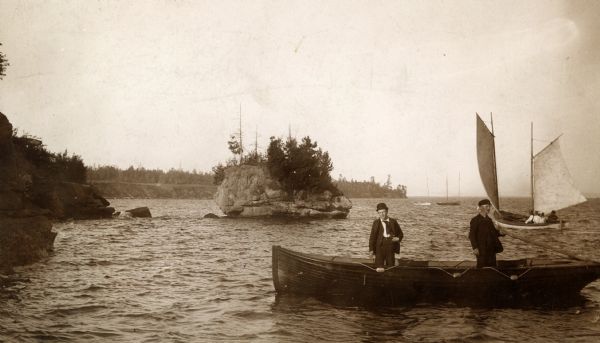 Two men standing in wooden rowboat along shore of Madeline Island.  There are sailboats in the background. Behind them is a large rock just off the shoreline, with trees growing on the side and top.