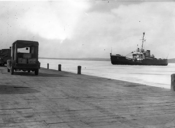 View from dock of the U.S. Coast Guard ship "Woodrush" arriving at the La Pointe dock to clear ice. Two trucks are on the dock, one with a piano.