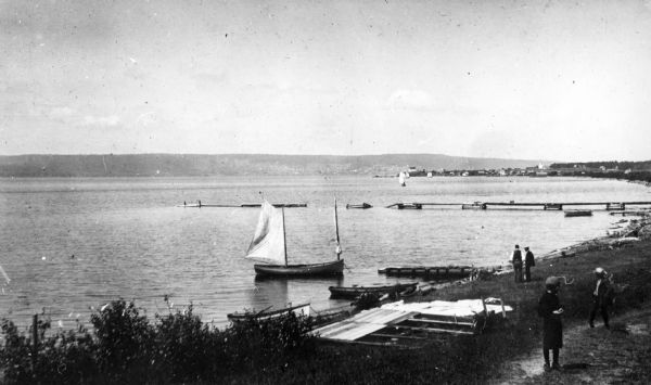 Elevated view of La Pointe and the shoreline from the Old Mission dock. Two boys and two men stand along the shoreline. The white building with the dark roof is the old school house before it was moved to the current location as the public library.