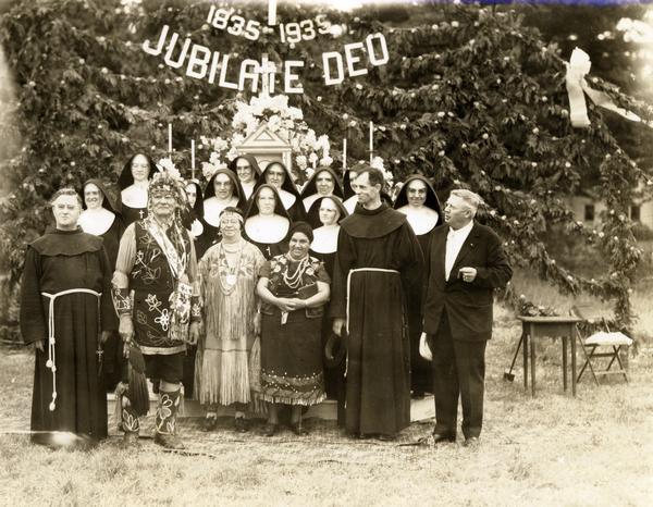 Father Gumbly and Sisters, with three Native Americans in ceremonial dress, standing outdoors in front of altar.