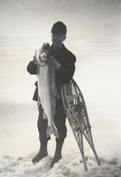 Fisherman standing on frozen Lake Superior holding a lake trout, leaning on snowshoes.
