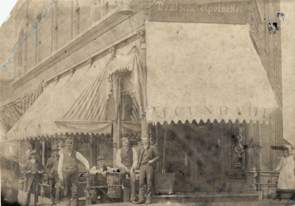 Customers gathered outside the J. Cunradi Drugstore.  A sign above the awning identifies the store as a <i>Deutsche Apotheke</i>, or German apothecary.