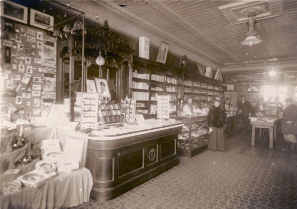 Colorful advertising displays line the counters of this turn-of-the- century drugstore, with rows of glass pharmaceutical bottles filling the shelves behind the counter.  A woman in a fur coat is served by a female clerk. One of the women is identified as Mrs. William H. Meyne.