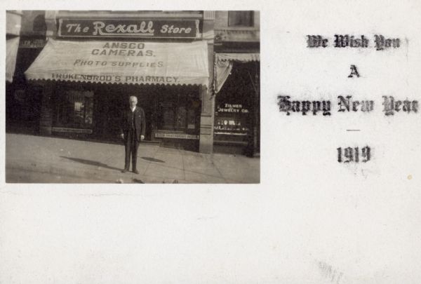W.F. Trukenbrod posing on the sidewalk underneath the awning of his store for this New Year's postcard. Text on right reads: "We Wish You A Happy New Year — 1919."