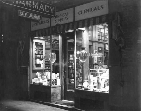Exterior view of the D.F. Jones' Pharmacy at night. 2 large showglobes are in the display windows.