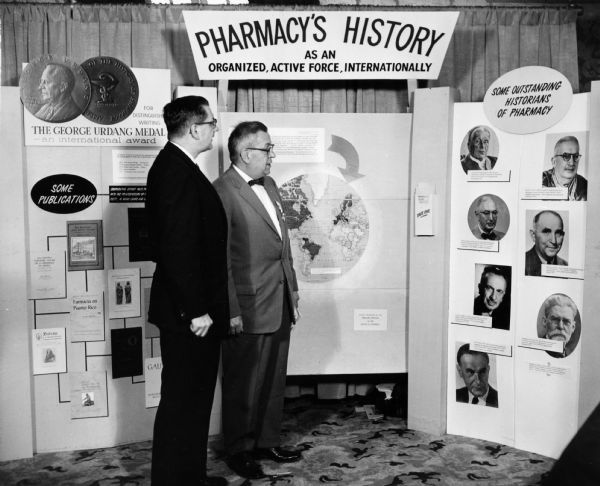 Pharmacist Donald Francke and a colleague examine a pictorial exhibit titled "Pharmacy's History: As an Organized, Active Force, Internationally".  The exhibit was organized by the American Institute for the History of Pharmacy.