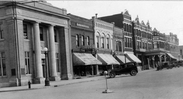 View of cars parked outside of several downtown businesses. Pictured from the left are a bank, Liese Drugstore, the Wright Bakery, two unidentified awnings, the Hotel Blodgett, and the Trio Theatre. 
