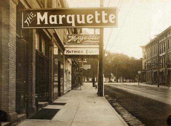 View up sidewalk with a view of storefronts lining King Street. The marquee of The Majestic is prominent in the foreground, as is The Marquette, and a drugstore further down the street. A man is looking out from behind a door on the far left. On the right side of the street is a sign for "The Variety Store, Marsh & Co." The Wisconsin State Capitol is in the background behind trees.