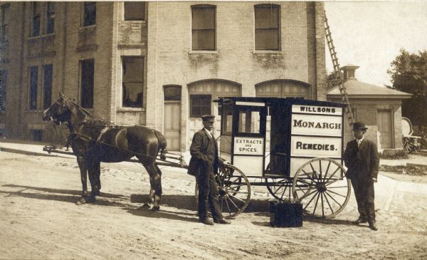 Two salesmen stand beside one of the Willson's Monarch Remedies horse-drawn wagons. Willson's sold patent medicines, spices, extracts, flavorings, and "toilet articles."