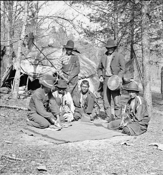 Six Ho-Chunk men are standing and kneeling around a blanket playing Wah-koo-chad-ah (Moccasin), a favorite game. A typical dwelling (chipoteke) is in the background.