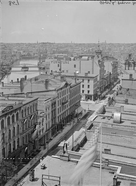 Milwaukee, north from Mitchell block. View of rooftops, buildings and bridge at distance on the left. American flag in the foreground.