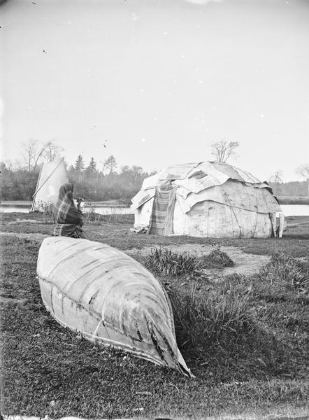 View towards a Ho-Chunk Indian wearing a blanket and holding a pipe standing behind a beached canoe. In the background are two typical dwellings (a chipoteke and a tepee or tipi) and beyond is a body of water.