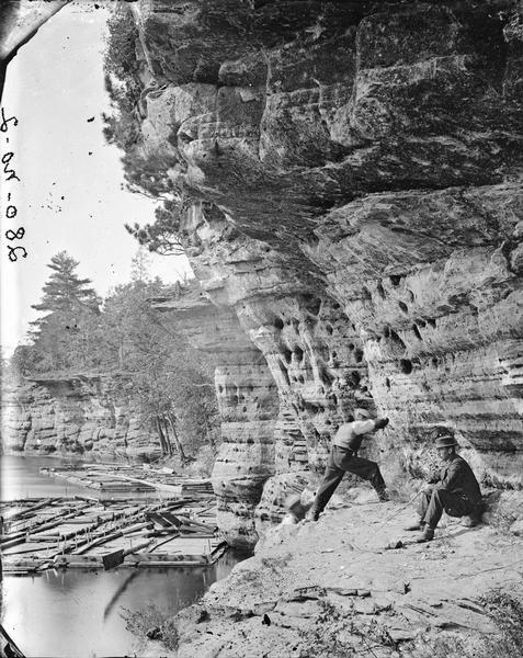 Two men at Swallows Nests, on sitting on the rocks at right, one leaning into the rock face. Two rafts are on the water under a cliff.