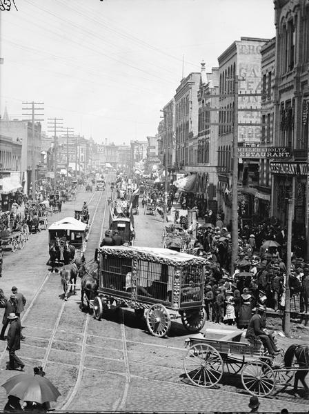 Circus parade at the corner of West Water and Grand Avenue. Crowds on the sidewalk and in horse-drawn vehicles are watching horse-drawn cages and other vehicles with flags.