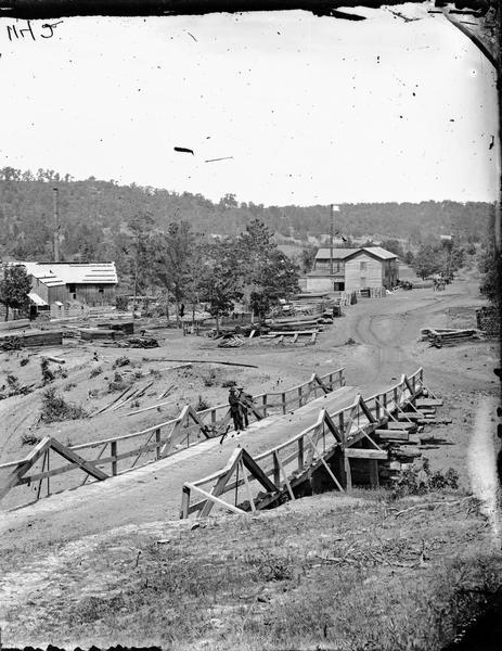 View of Kilbourn, with Superior Street bridge in Happy Hollow. Two men are standing on a bridge. Buildings and stacked lumber are in the background.