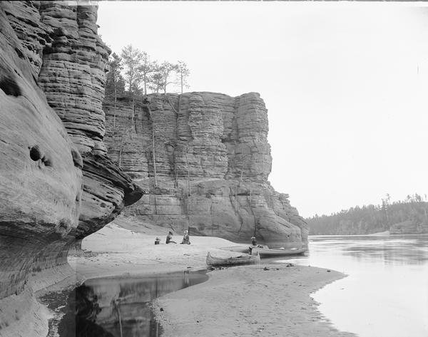 At the foot of High Rock (north face). Two women are sitting on the shore. On the right is a third woman sitting on a canoe at the edge of the river.