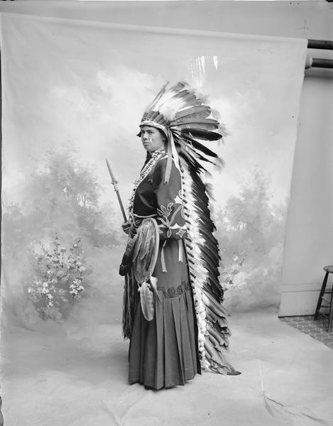 Full-length studio portrait in front of a painted backdrop of an unidentified person in Native American costume.