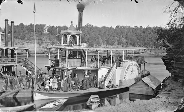 Steamboat <i>Alexander Mitchell</i> and edge of second unidentified steamboat at shore, with passengers posed standing on the lower deck and sitting on the upper deck. Rowboats on shore.