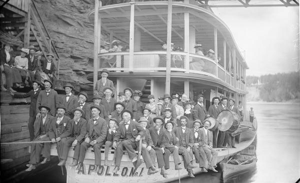 Passengers posing on bow of "Apollo No. 1" steamboat. Two men with drums are on the right. Edge of bridge a is at top.