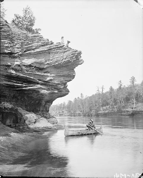 Up the river along the shoreline, opposite from Robinson's Landing. There is a man in a canoe in the river near the shoreline. Two women are on the protruding rock ledge above.