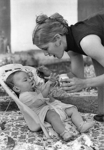 A young mother feeds her infant baby food outdoors.